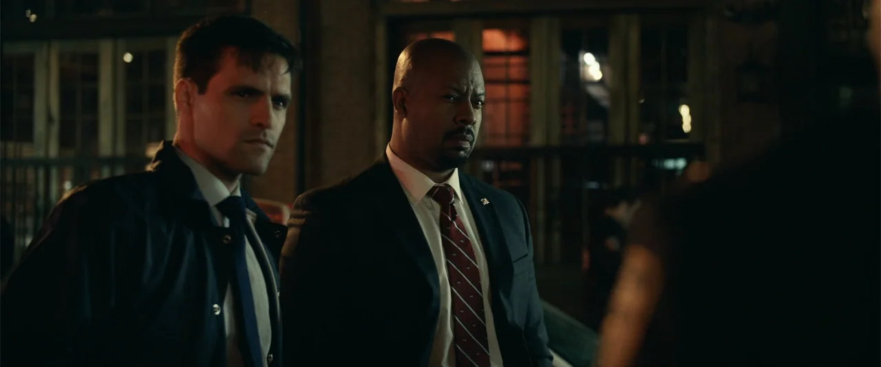 21 Bridges (2019) movie still: medium — A night city street features two men in business suits standing in the foreground. They both look forward…