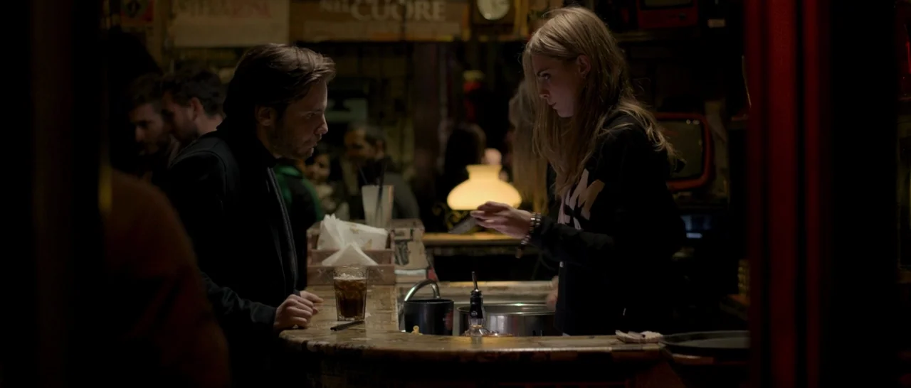The Face of an Angel (2014) movie still: medium — A dimly lit bar with wooden surfaces and an illuminated lamp in the background. A man stands at…