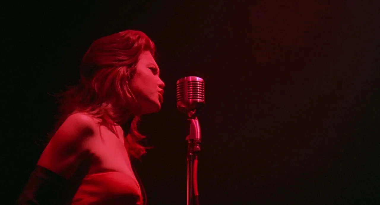 Streets of Fire (1984) movie still: medium — A dark, empty nightclub stage features a single microphone stand under a red spotlight. A woman…