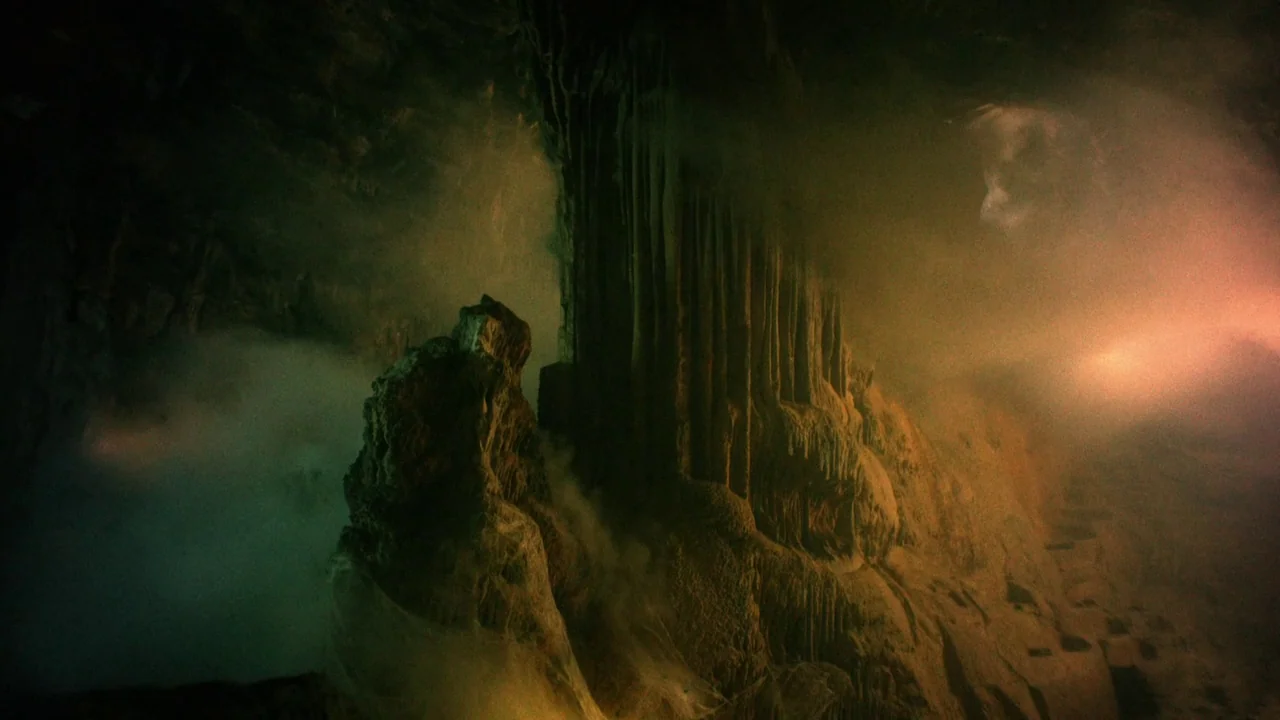 The Field Guide to Evil (2018) movie still: establishing — A dark subterranean cavern contains jagged limestone rock formations and thick drifting…