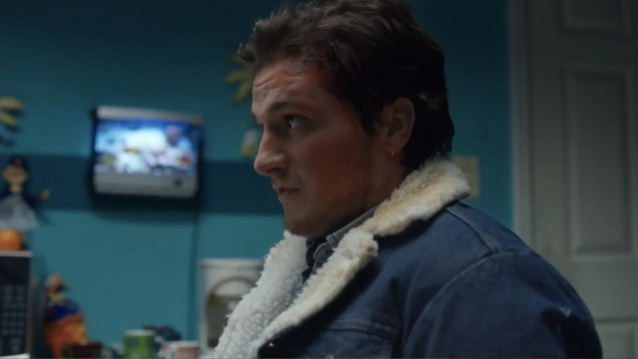 Burn (2019) movie still: medium — A residential kitchen features blue walls and a small television screen mounted above cabinets. A man stands in…