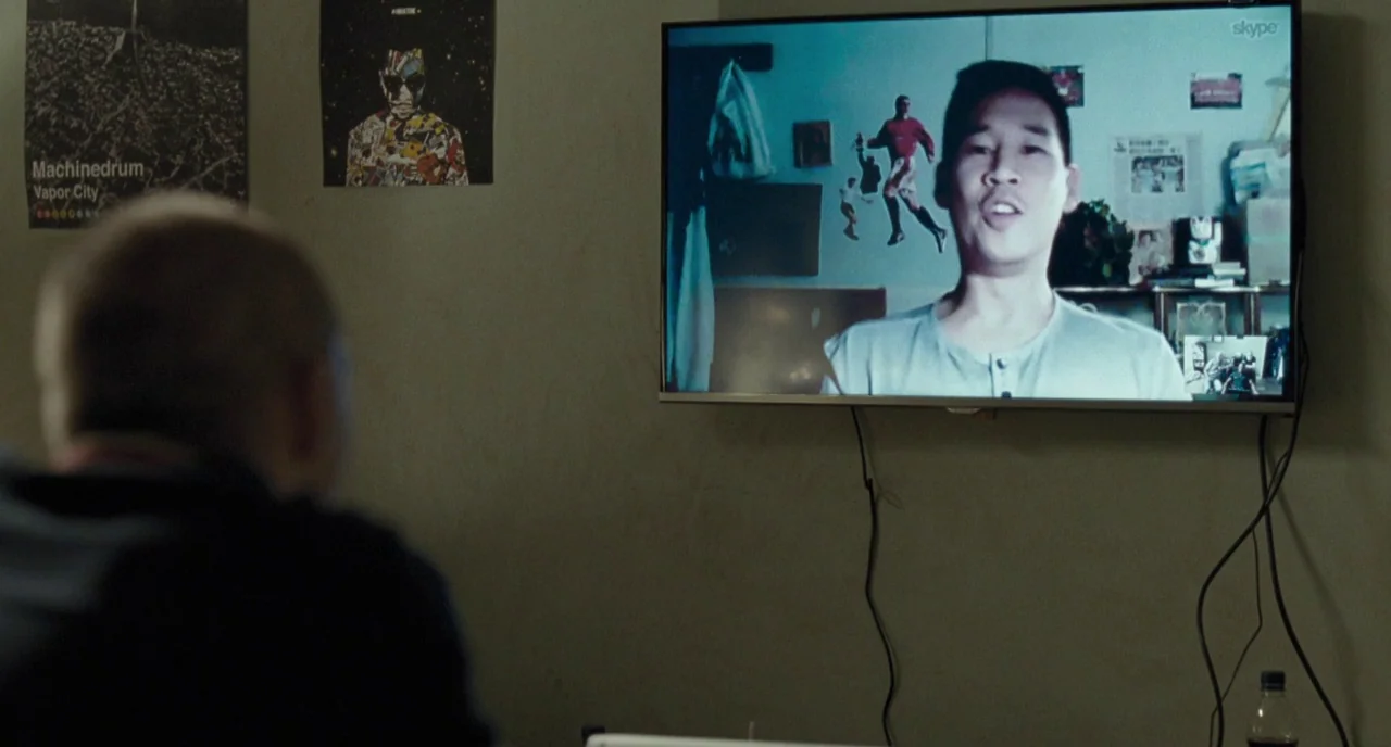 I, Daniel Blake (2016) movie still: medium — A dimly lit room features a large television displaying a Skype video call. A man on screen talks while…