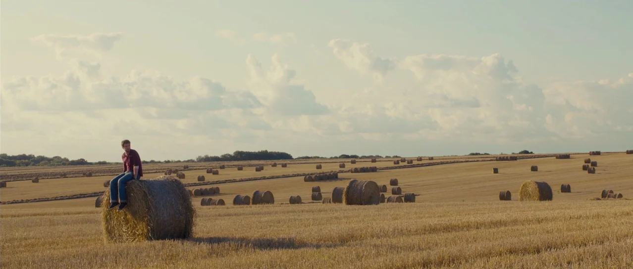 Road Games (2015) movie still: wide — A vast harvested wheat field filled with scattered round hay bales under a sky. A man sits alone on top of the…