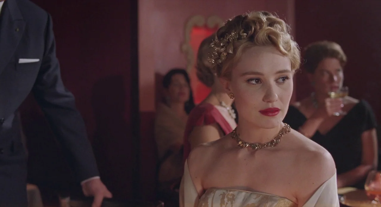 Populaire (2012) movie still: medium — A dimly lit room with maroon walls contains guests seated at round tables. A blonde woman in a gold dress…