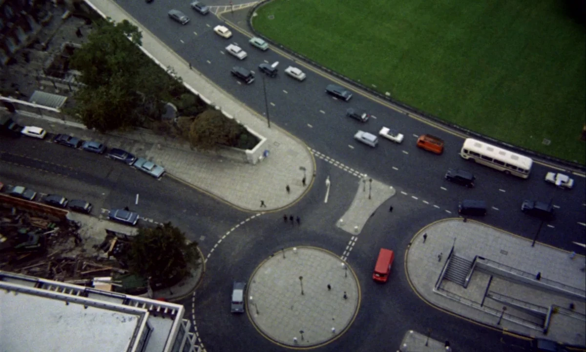 Wonderwall (1968) movie still: establishing — A high-angle view looks down upon a concrete roundabout and intersecting city roads. Small cars, a…