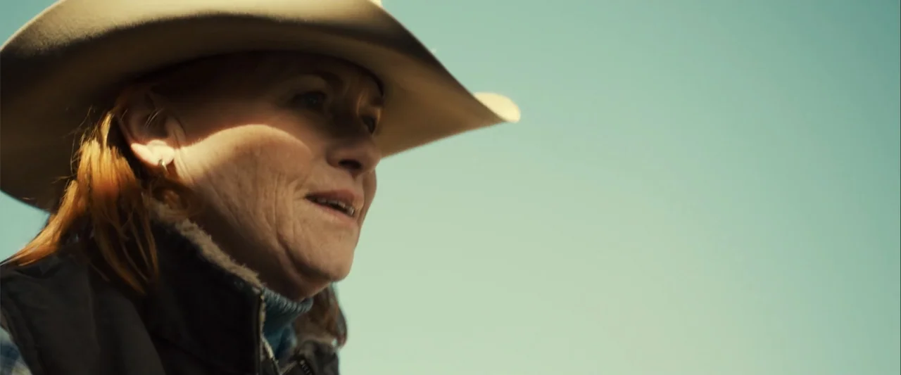 Frontera (2014) movie still: closeup — An outdoor location with a clear, light blue sky in the background. A woman in a cowboy hat looks off-camera…