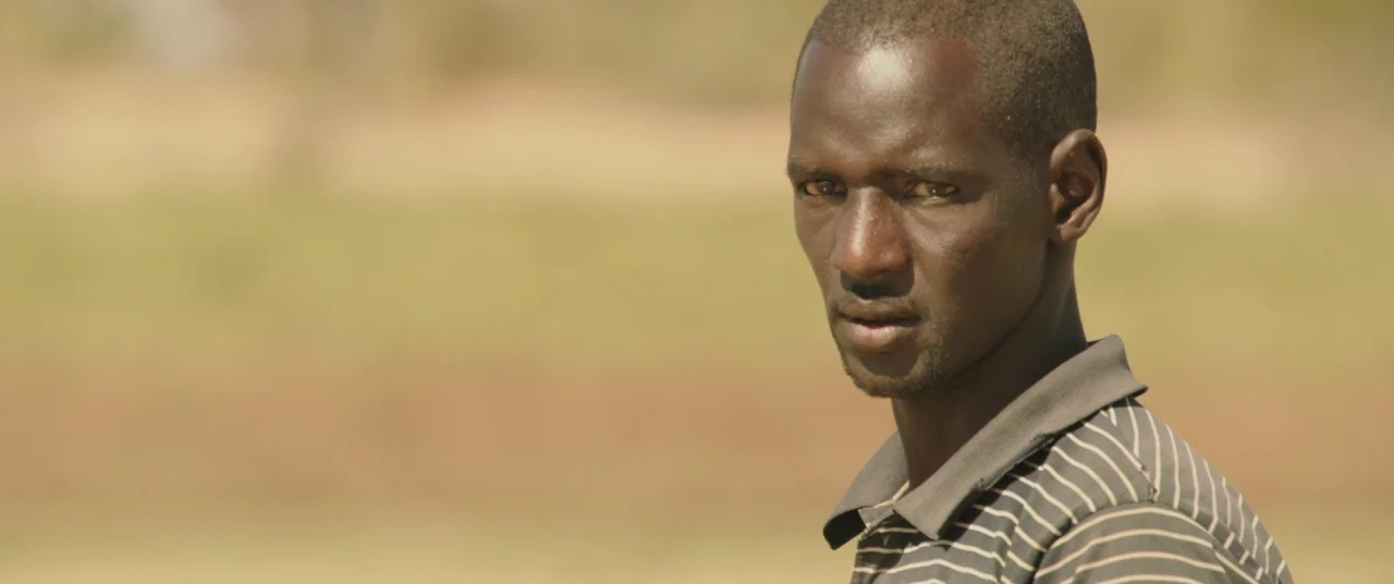 Timbuktu (2014) movie still: closeup — An open rural landscape with blurred tan and green vegetation in the background. A young man turns his head…