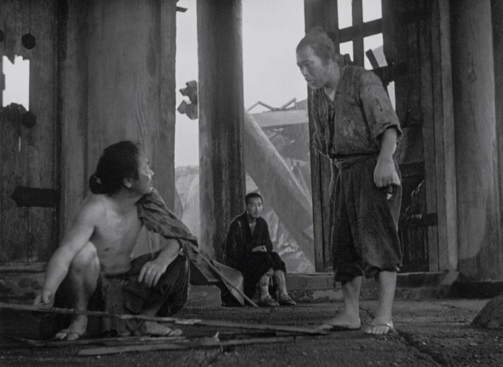 Rashomon (1950) movie still: medium — A dilapidated wooden gatehouse with large columns shows remnants of heavy damage and decay. Two men engage in…
