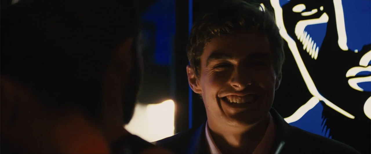 Unfinished Business (2015) movie still: closeup — A dimly lit interior bar setting features a glowing blue sign in the background. A young man in a…
