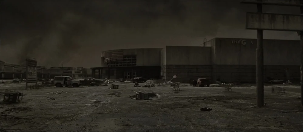 The Road (2009) movie still: establishing — An abandoned suburban shopping mall parking lot features cracked asphalt and scattered shopping carts.…
