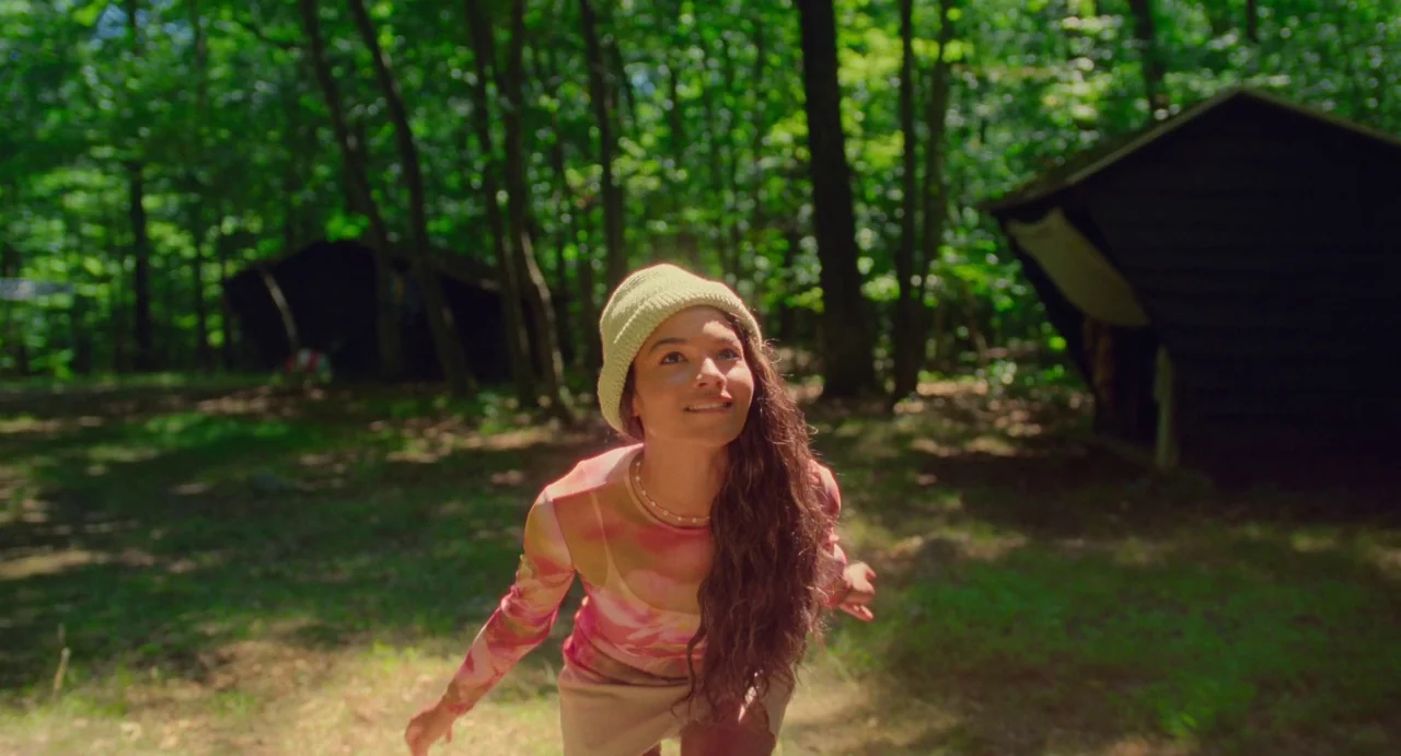 I Saw The TV Glow (2024) movie still: medium — A sunny campsite in a deciduous forest with wooden shelter structures in the background. A woman in a…