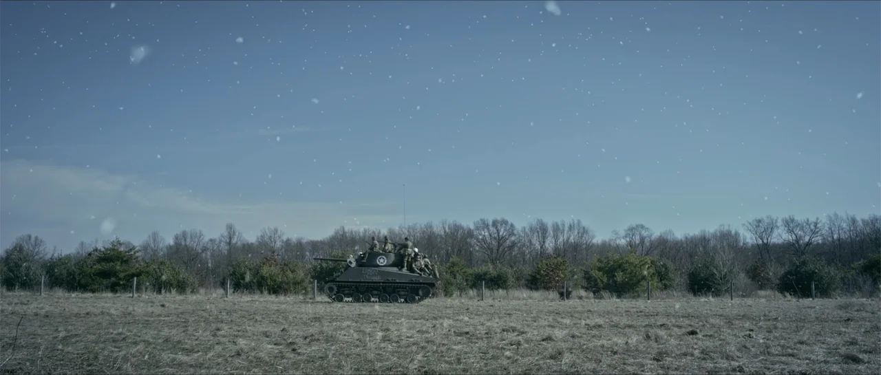 Battle of the Bulge (1965) movie still: wide — A dark green tank traverses a vast, dry field surrounded by bare winter trees. Light snow falls…