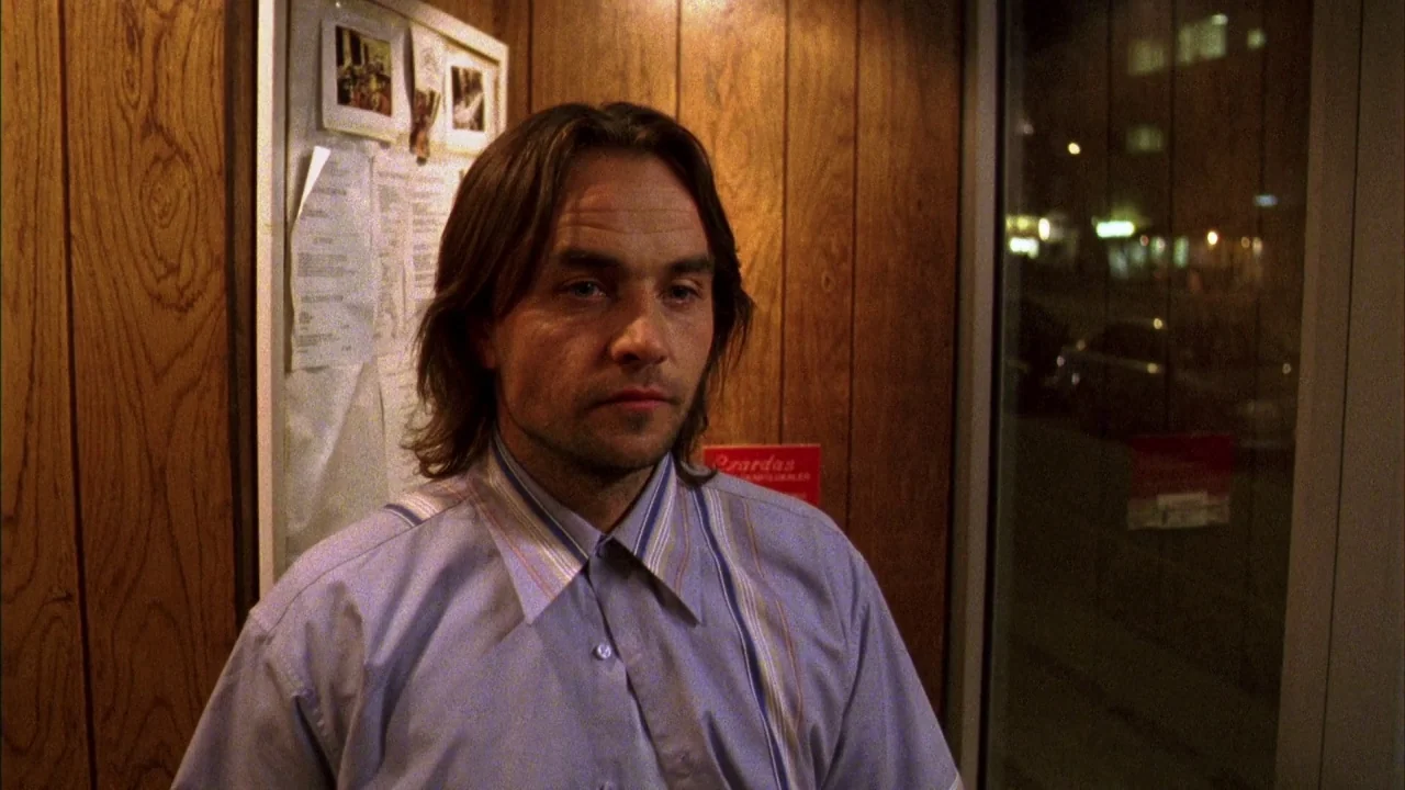 Pusher III (2004) movie still: medium — A room featuring vertical wood-paneled walls and a glass window overlooking a street at night. A man with…