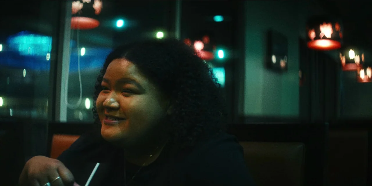 Spermworld (2024) movie still: medium — A dark booth inside a restaurant features low-light conditions with colored neon signage visible. A young…