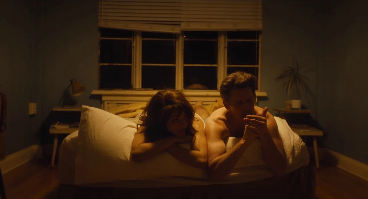 Born To Be Blue (2015) movie still: medium — A dimly lit room features a bed positioned against a window with closed blinds. A man and a woman rest…