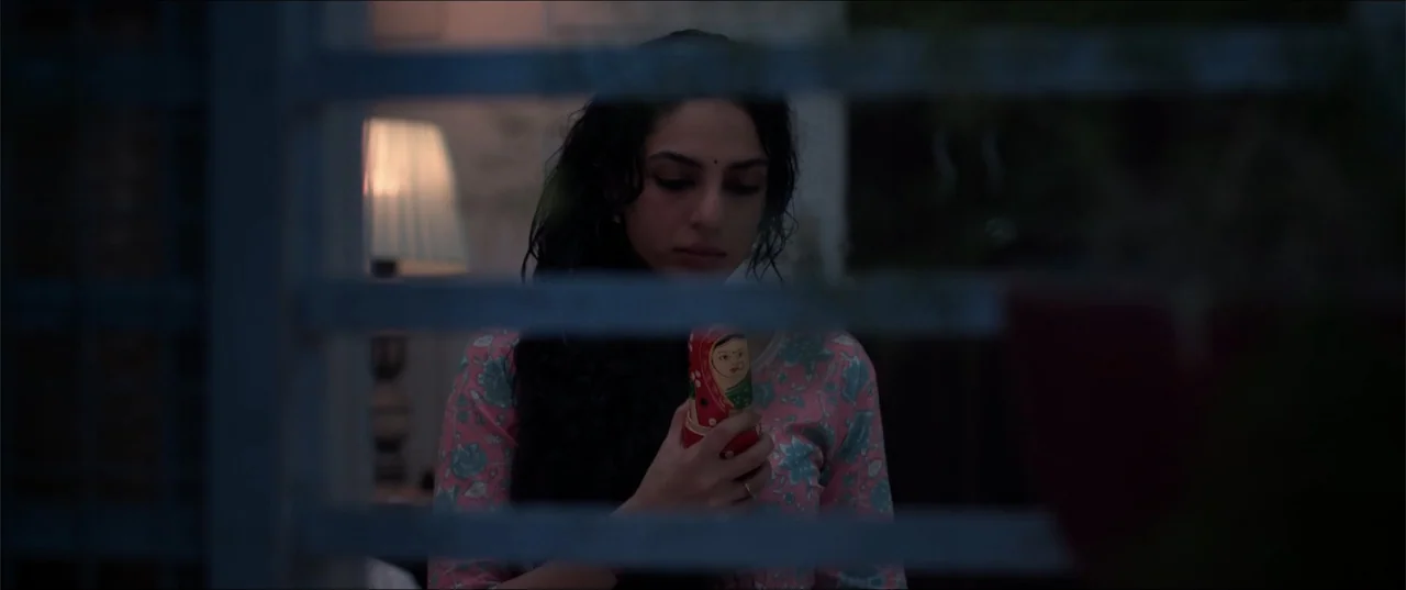 Cheekatilo (2026) movie still: medium — A dim room interior with a woman standing behind a slatted window frame. A woman with dark hair holds a…