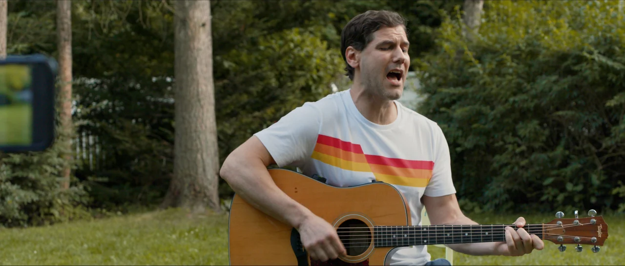 Songs For A Sloth (2021) movie still: medium — A grassy outdoor area with mature trees visible in the soft-focus background. A man plays an acoustic…
