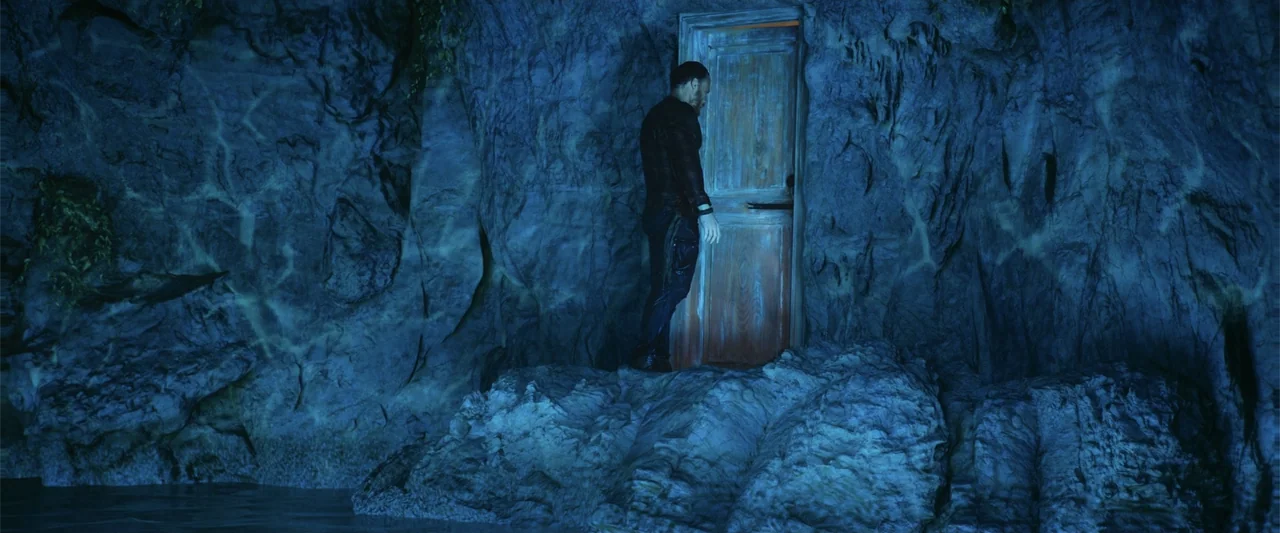 Blue Cave (2024) movie still: fullbody — A natural cave interior features rugged stone walls and a lone wooden door. A man stands motionless before…