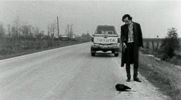 Roadkill (1989) movie still: fullbody — A desolate rural asphalt road runs through a sparse, leafless landscape under overcast skies. A man stands…