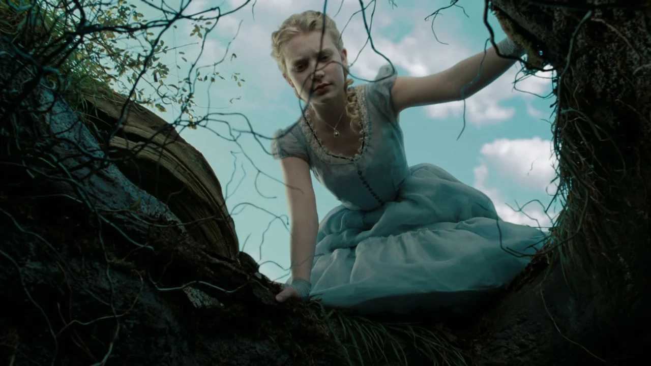 Alice in Wonderland (2010) movie still: medium — A girl looks down into a dark, textured opening within a gnarled tree trunk. She is framed from…