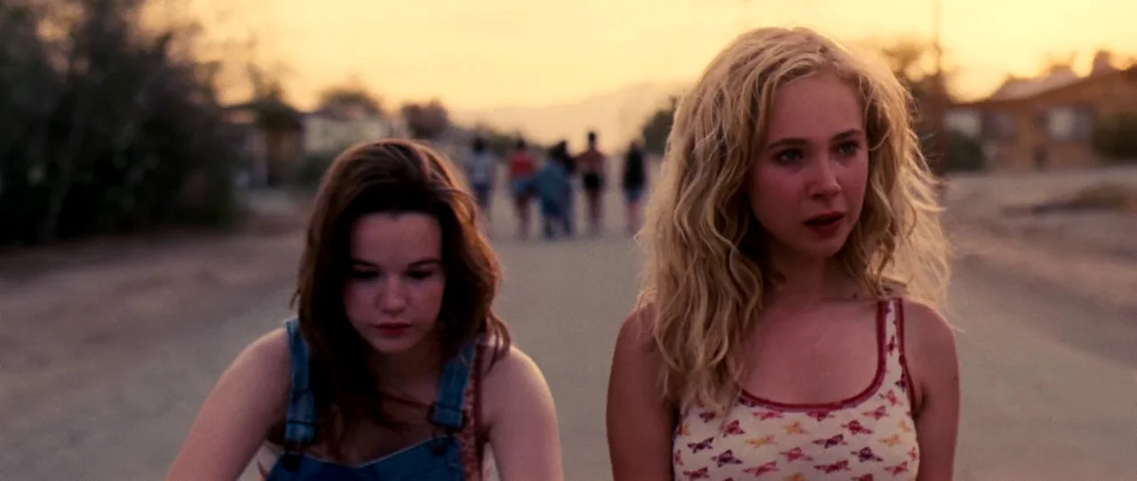 Little Birds (2011) movie still: medium — A suburban road stretches into the distance under the warm glow of sunset. Two teenage girls ride bicycles…