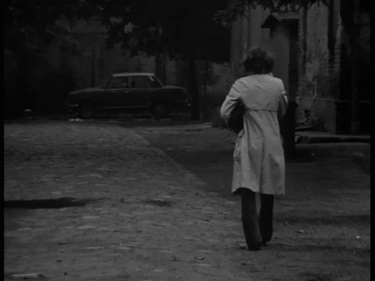 Family Nest (1979) movie still: fullbody — An empty cobblestone courtyard features a parked vintage sedan near dark trees and buildings. A woman in…