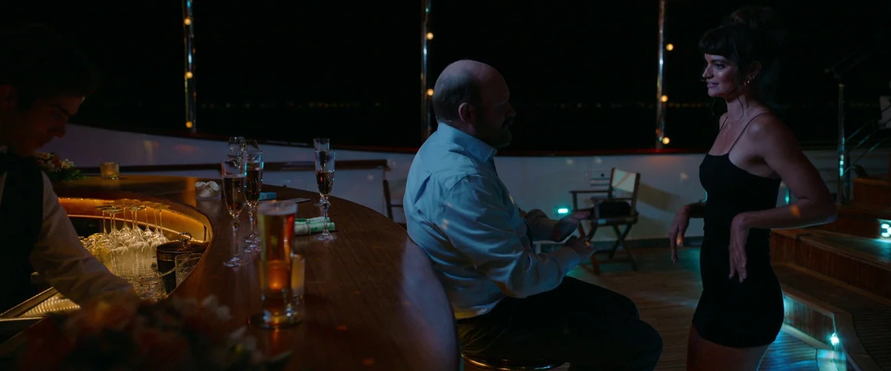 Triangle of Sadness (2022) movie still: medium — A dark outdoor yacht deck at night features a curved wooden bar counter. A man sits at the bar…