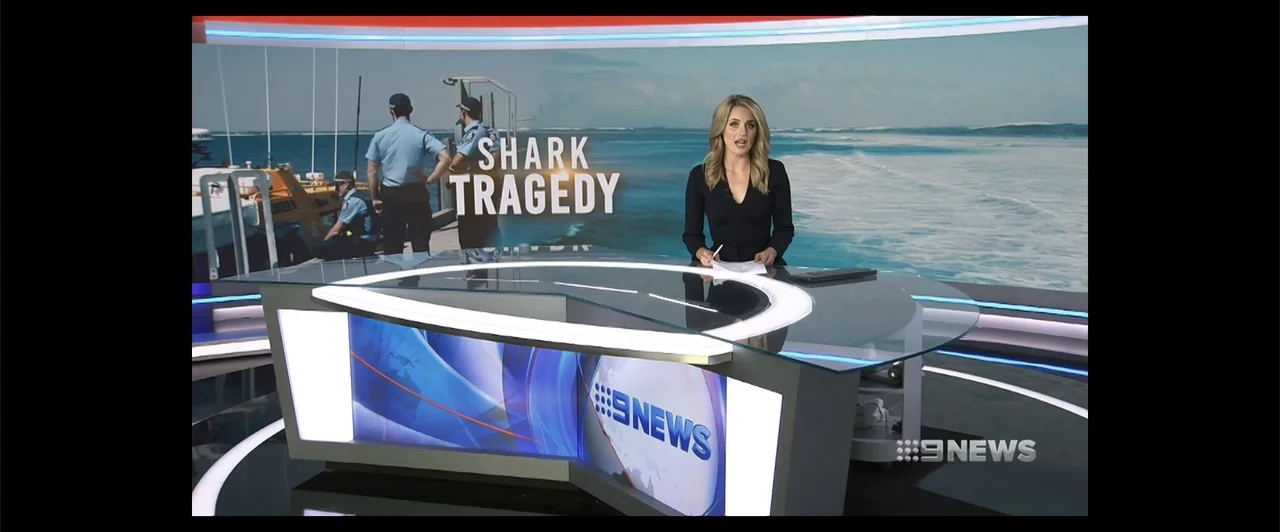 Lair of the Great White (2024) movie still: medium — A modern news studio desk sits before a large digital screen background. A female news anchor…