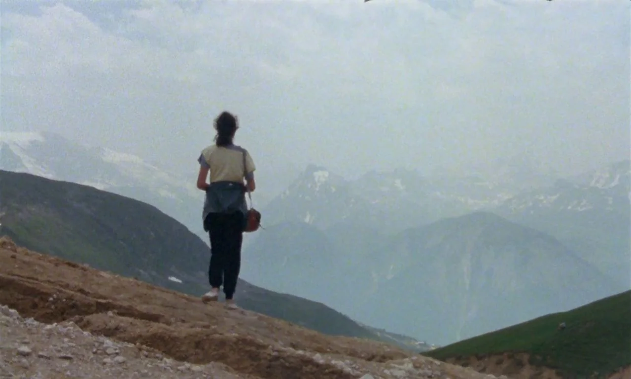 The Green Ray (1986) movie still: fullbody — A person walks on a dirt trail on the side of a mountain. Distant snowy peaks rise through low clouds…