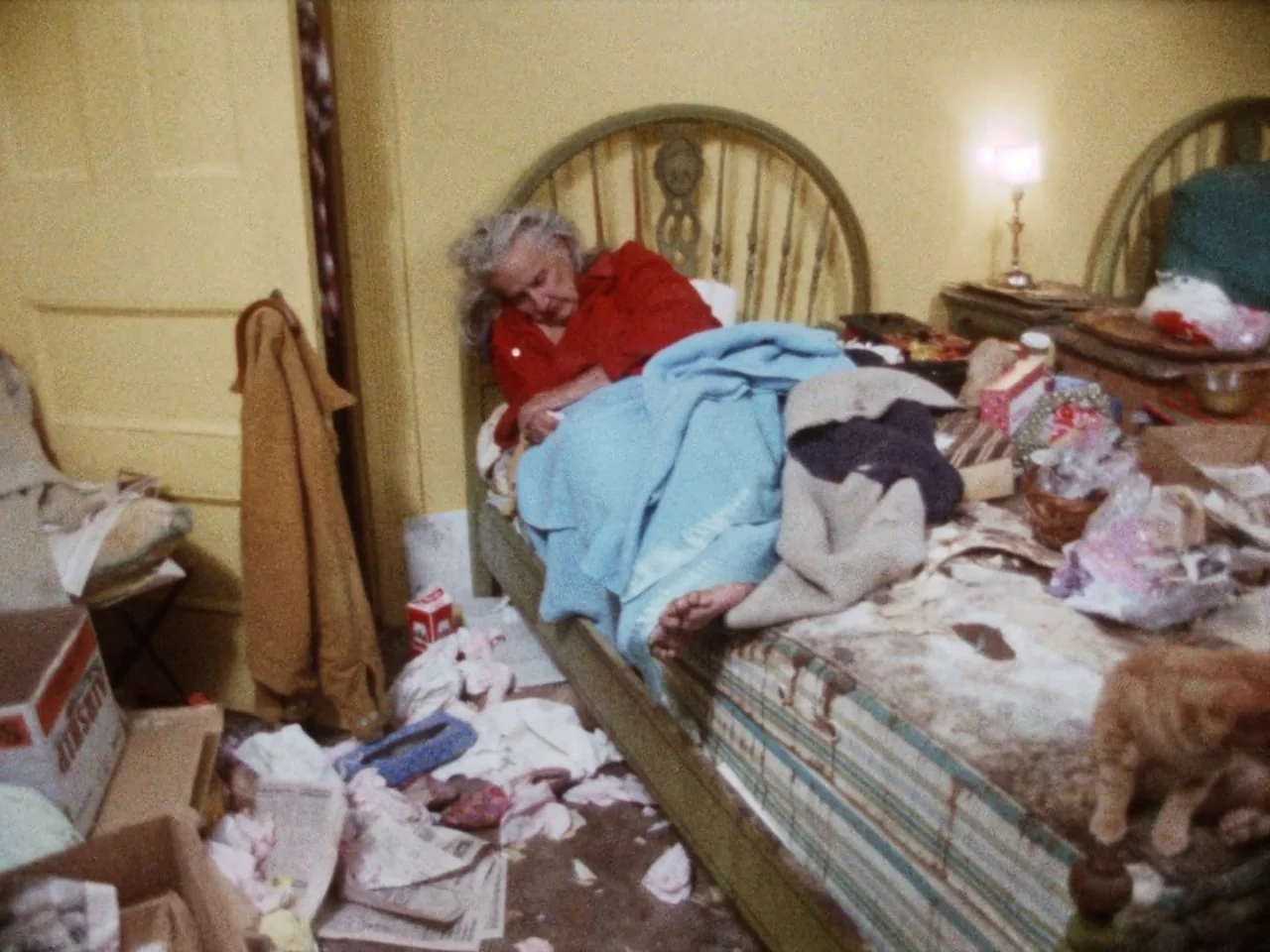 Grey Gardens (1975) movie still: medium — A cluttered room features a bed covered in blankets and various personal items. An elderly woman lies on…