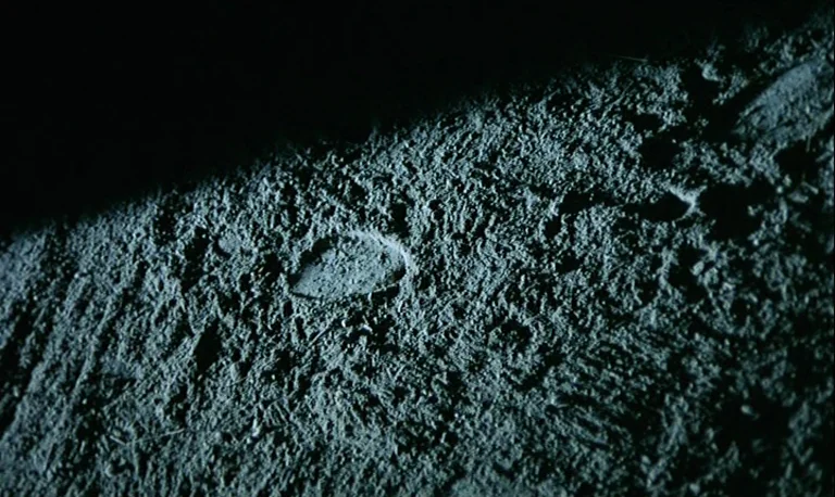 The Rose King (1986) movie still: closeup — A patch of uneven, dark soil displays a clear indentation of a footprint. The texture of the ground is…