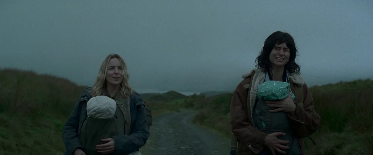 The End We Start From (2023) movie still: fullbody — Two women walk along a gravel road in a misty, highland environment. Each woman wears a baby…