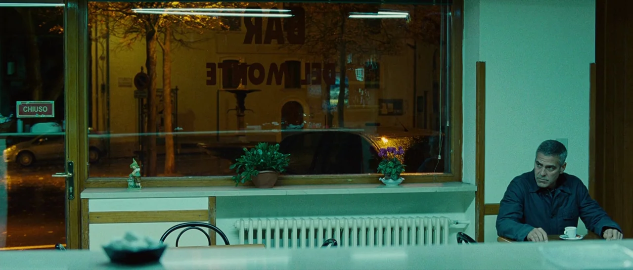 The American (2010) movie still: wide — A quiet, dimly lit cafe at night with green-tinted fluorescent lighting. George Clooney sits alone at a…