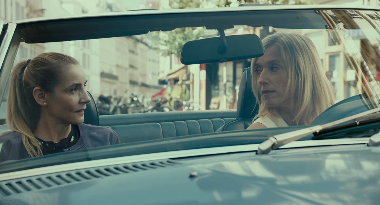 Last Summer (2023) movie still: medium — A light blue vintage convertible car sits on a city street with blurred urban buildings. Two middle-aged…