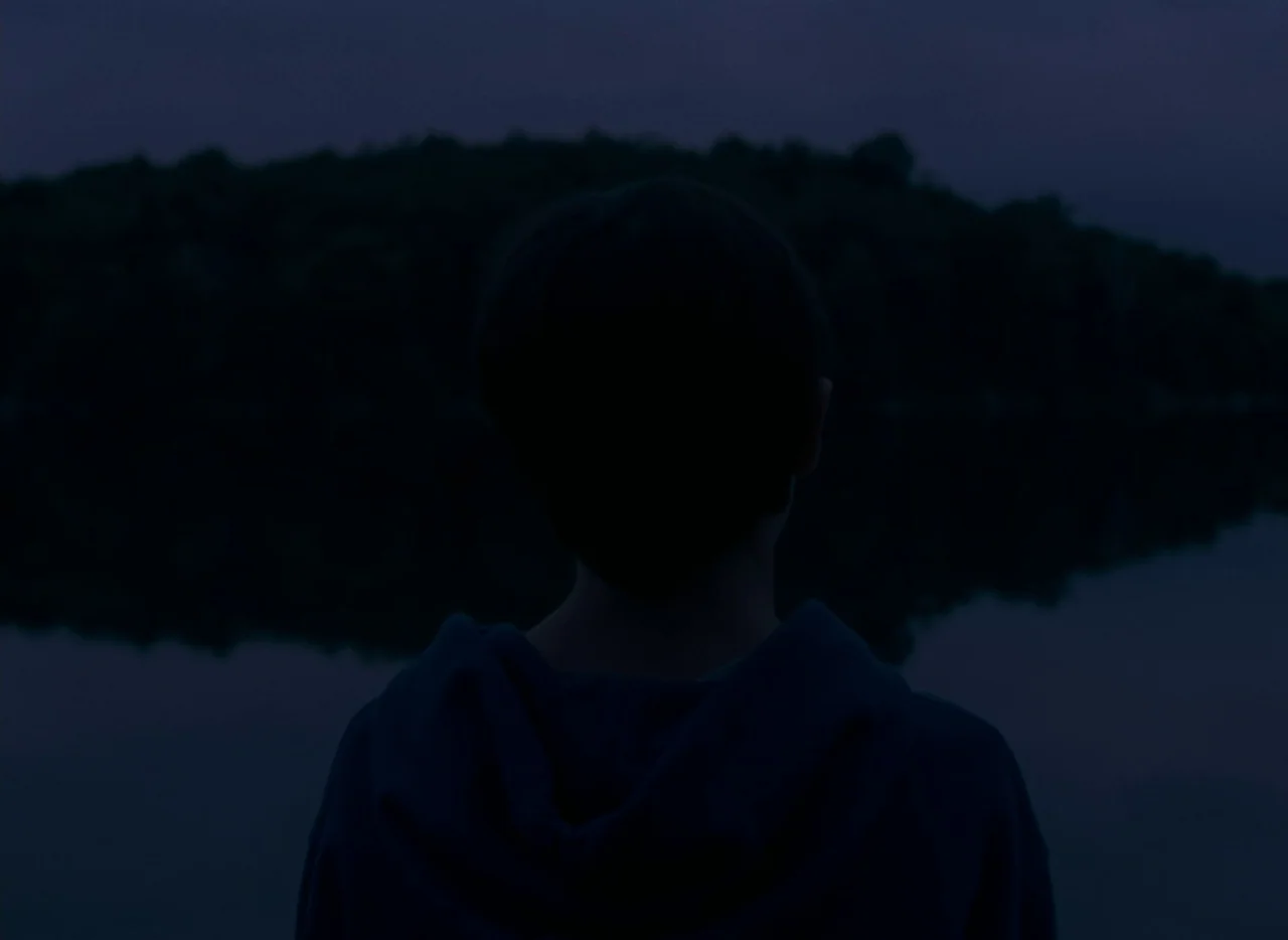 Falcon Lake (2022) movie still: medium — A dark body of water is bordered by a tree-lined bank under twilight skies. A person in a hooded sweatshirt…