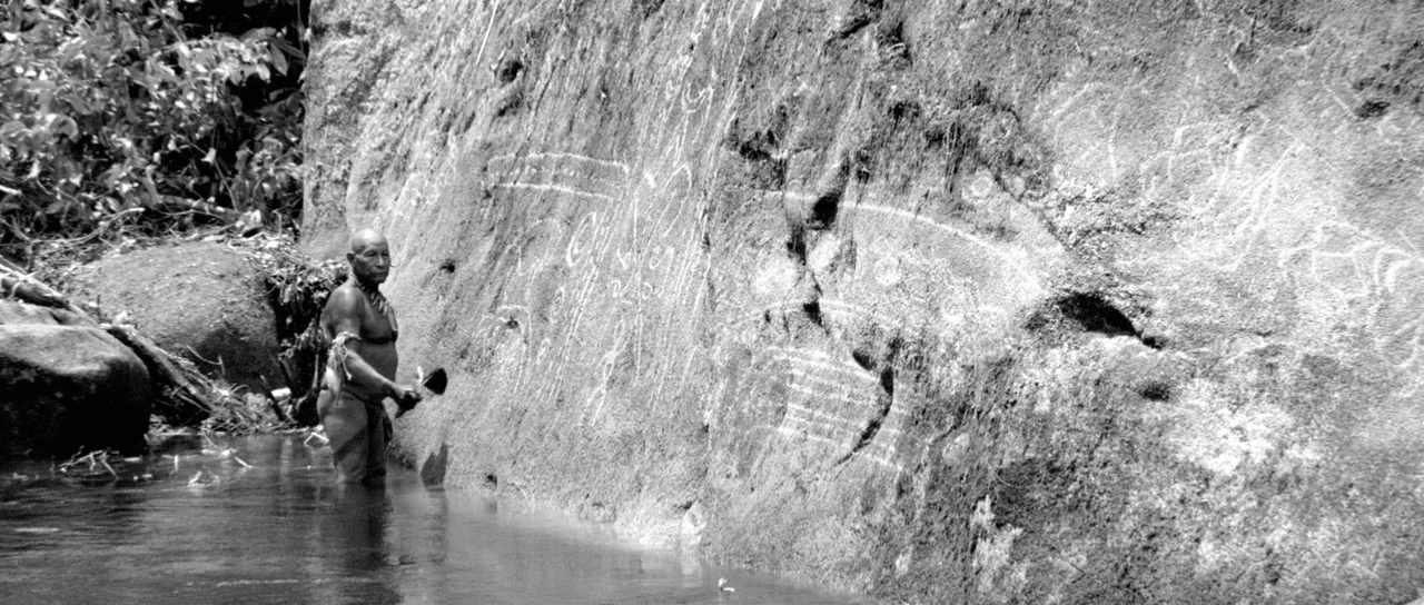 Embrace Of The Serpent (2015) movie still: wide — An elderly indigenous man stands in a shallow stream beside a large rock wall. The stone surface…