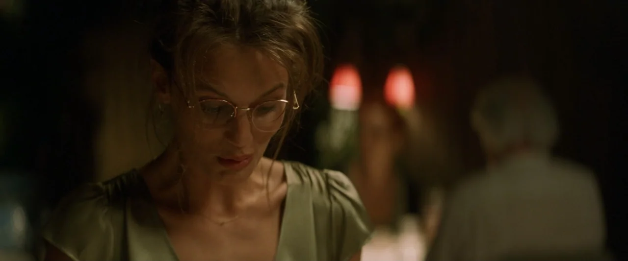Masquerade (2022) movie still: closeup — A dark indoor space features a woman in an olive blouse dining. The subject looks down at a table while…