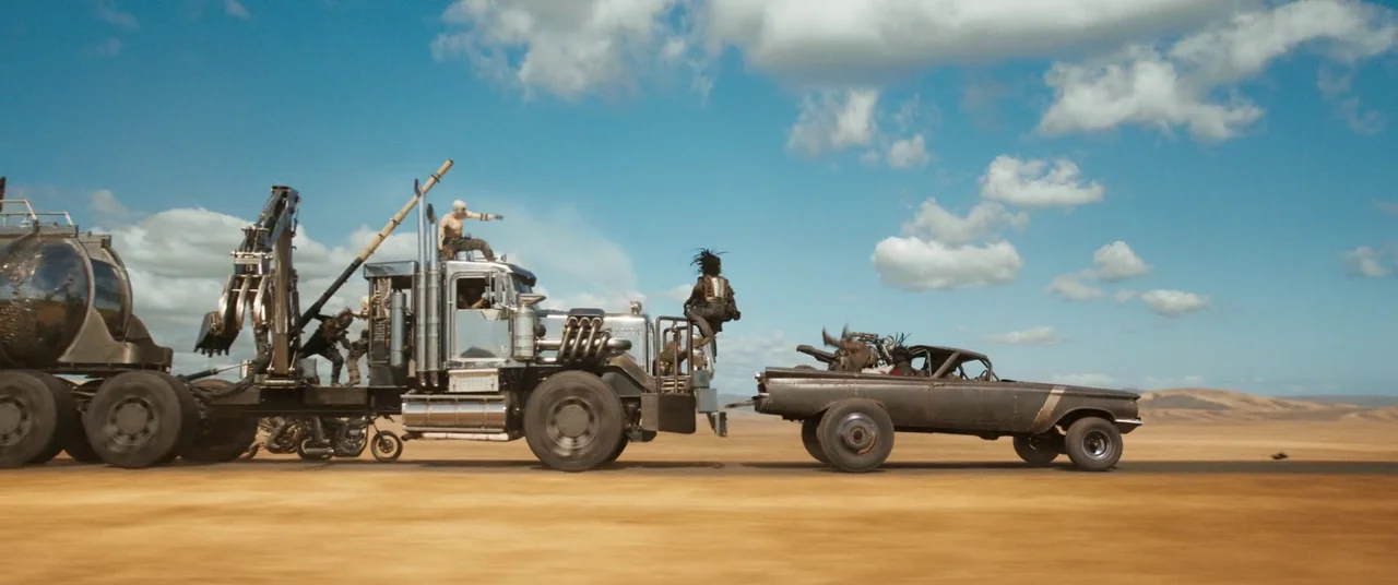 Furiosa: A Mad Max Saga (2024) movie still: wide — A vast, flat desert landscape stretches under a bright blue sky with scattered clouds. A modified…