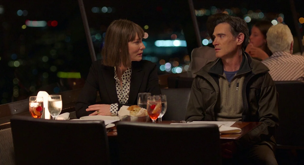 Where’d You Go Bernadette (2019) movie still: medium — A dimly lit upscale restaurant interior overlooks a distant city skyline at night. A man and…