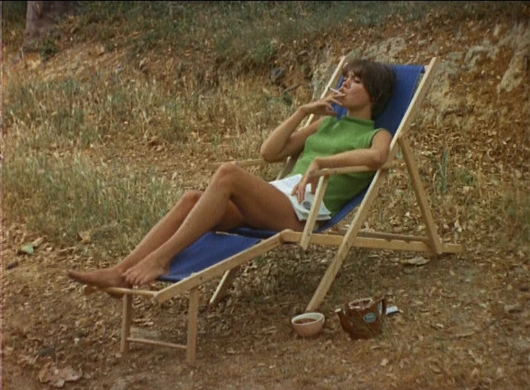 La Collectionneuse (1967) movie still: fullbody — A woman relaxes in a blue wooden deck chair on dry, patchy grass. She holds a cigarette in one…