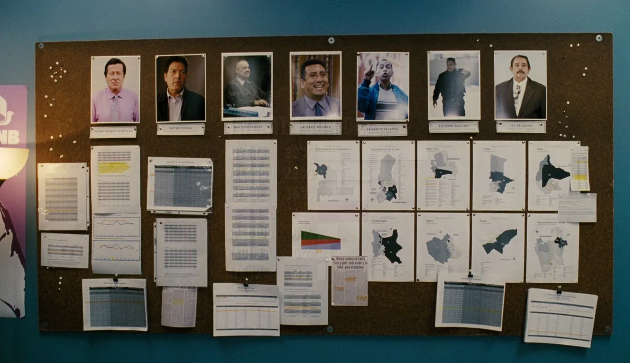 Our Brand is Crisis (2015) movie still: wide — A dark brown corkboard is mounted on a solid blue wall. Numerous paper documents, maps, and portrait…