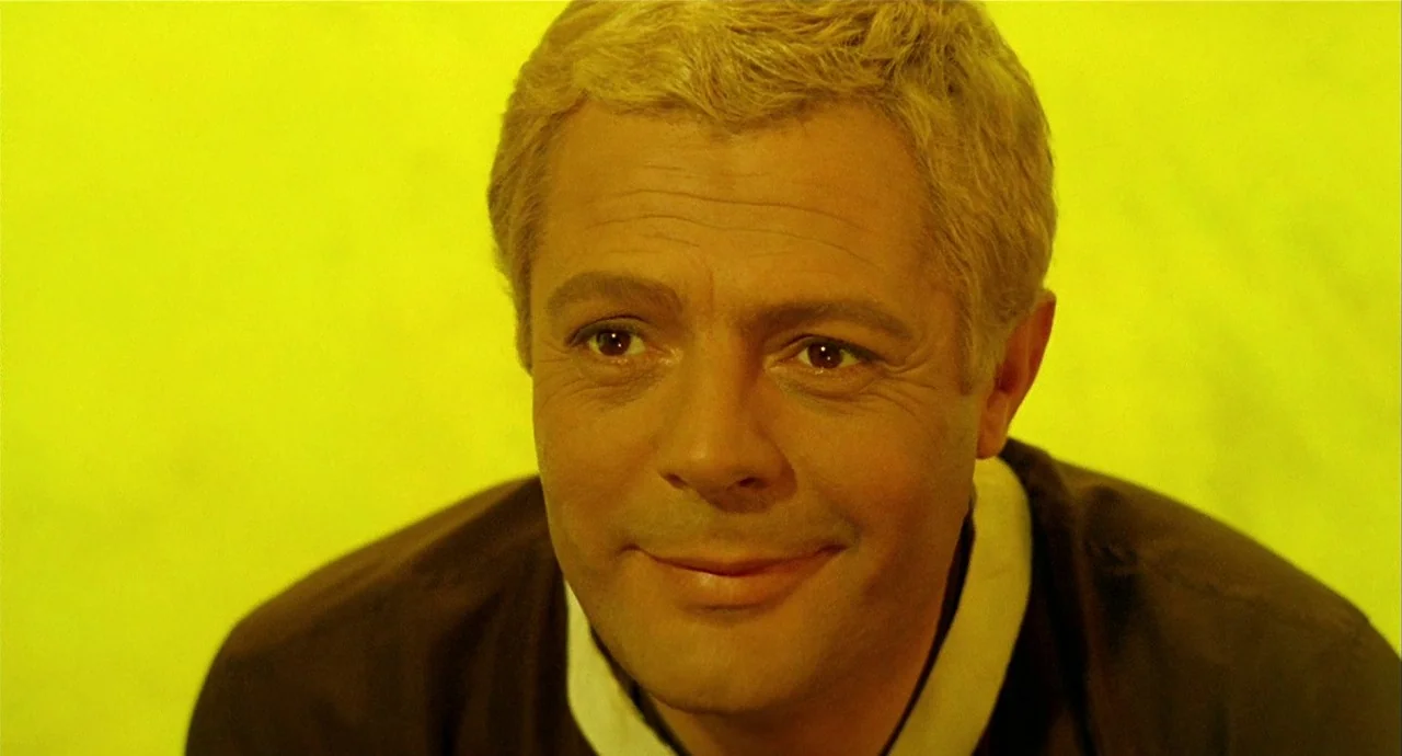 The 10th Victim (1965) movie still: closeup — A man is positioned against a vibrant, solid yellow screen that fills the frame. He looks forward with…