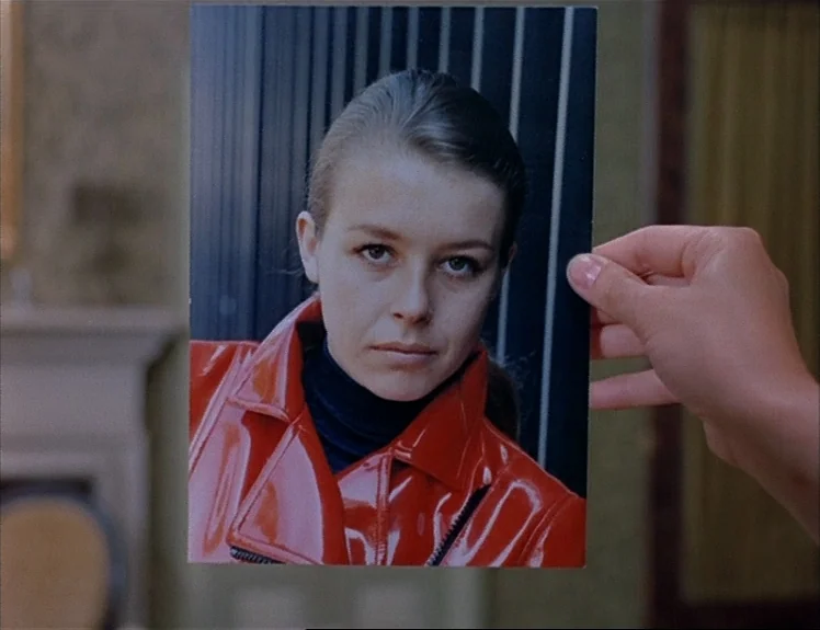 Claire’s Knee (1970) movie still: closeup — A hand holds a color photograph depicting a woman in a red jacket. The background is a soft-focus…