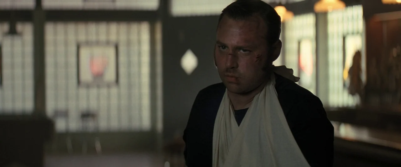 The Bikeriders (2023) movie still: medium — A dimly lit room features walls constructed of translucent glass block tiles. A man with a bandaged arm…