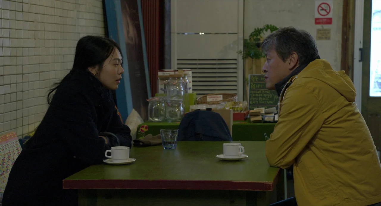On The Beach At Night Alone (2017) movie still: medium — A quiet indoor space features a small table with two white ceramic cups. Two people sit…