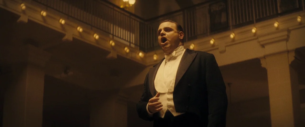The Immigrant (2013) movie still: medium — An opulent ballroom features an elevated mezzanine lined with circular golden light fixtures. A man in a…