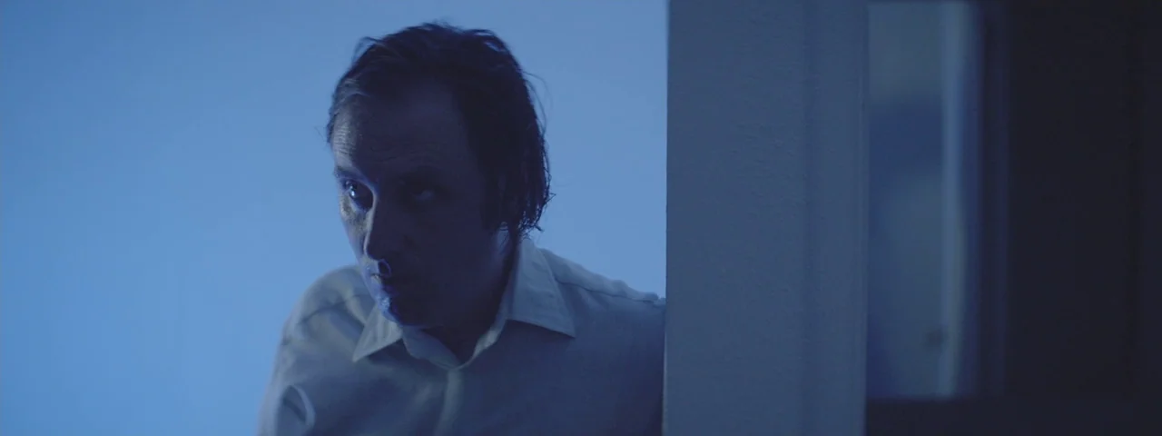 Entertainment (2015) movie still: medium — A man in a white shirt peeks around a wall in a darkened space. He stares intently forward while…
