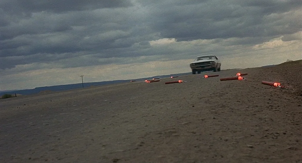 Vanishing Point (1971) movie still: wide — An asphalt road stretches into the distance under a heavy, overcast sky. Several illuminated red flares…