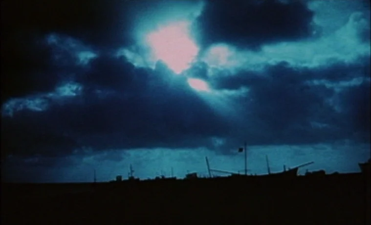 The Garden (1990) movie still: establishing — A dark harbor scene at night featuring silhouettes of docked boats under cloudy skies. Moonlight…