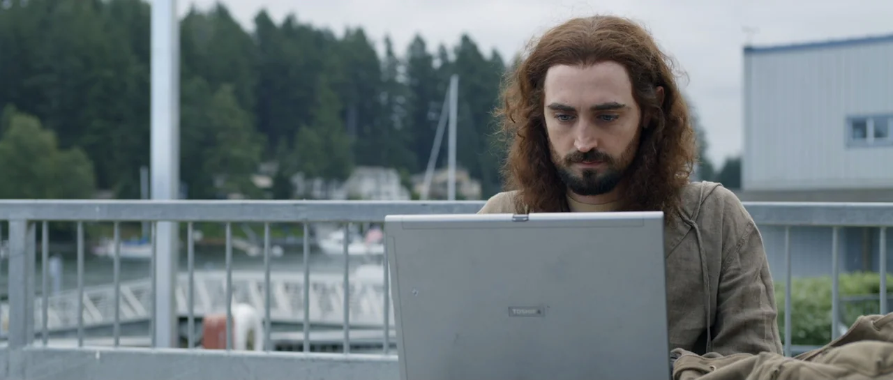 La presencia (2012) movie still: medium — A man sits at a railing overlooking a harbor with trees in the background. He is using a silver laptop…